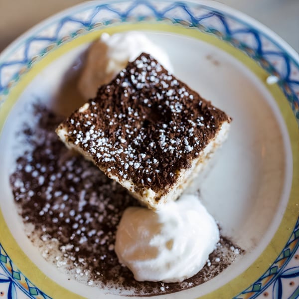 A slice of tiramisu with cocoa powder and powdered sugar, served with whipped cream on a decorative plate.