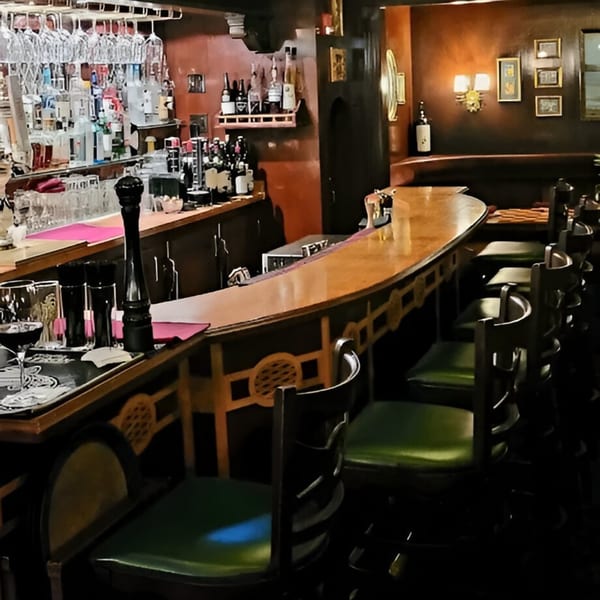 A dimly lit, cozy bar at Italian Kitchen with a wooden counter and green-seated stools.