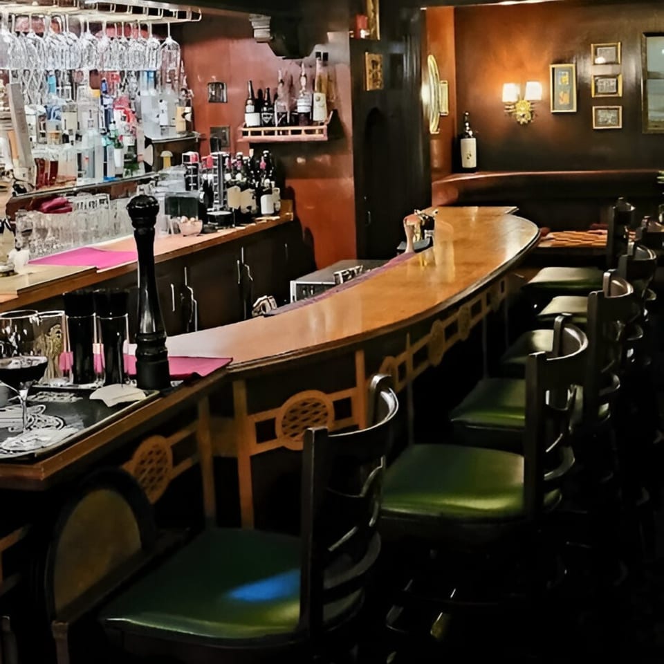 A dimly lit, cozy bar at Italian Kitchen with a wooden counter and green-seated stools.