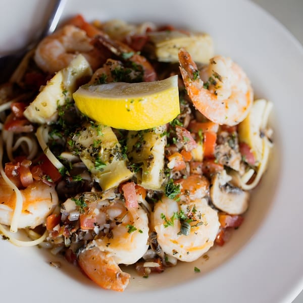 Shrimp scampi with linguine, artichoke hearts, diced tomatoes, mushrooms, and a lemon wedge.